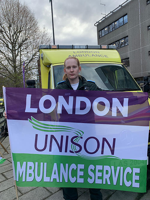 Trainee paramedic technician Eden at the Waterloo picket, in front of an ambulance and with a Greater London UNISON Ambulance Service flag