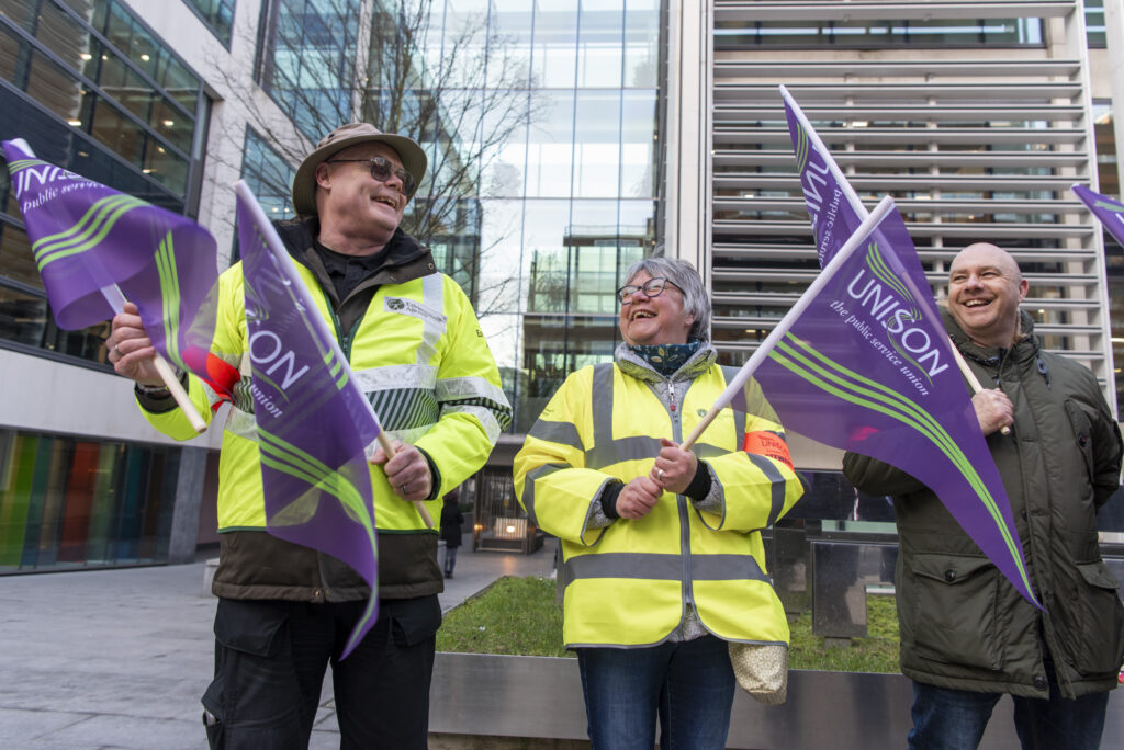 Two people in High vis tops wave UNISON flags on an EA picket line